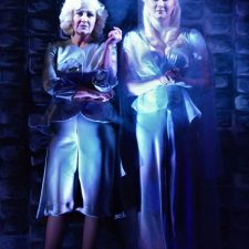 Elvira and Ruth standing in dim blue light with fog behind them